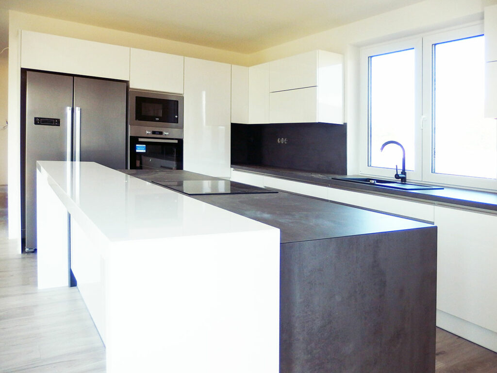 Kuchyň do L s ostrůvkem v bílé lesklé barvě a šedou pracovní deskou.