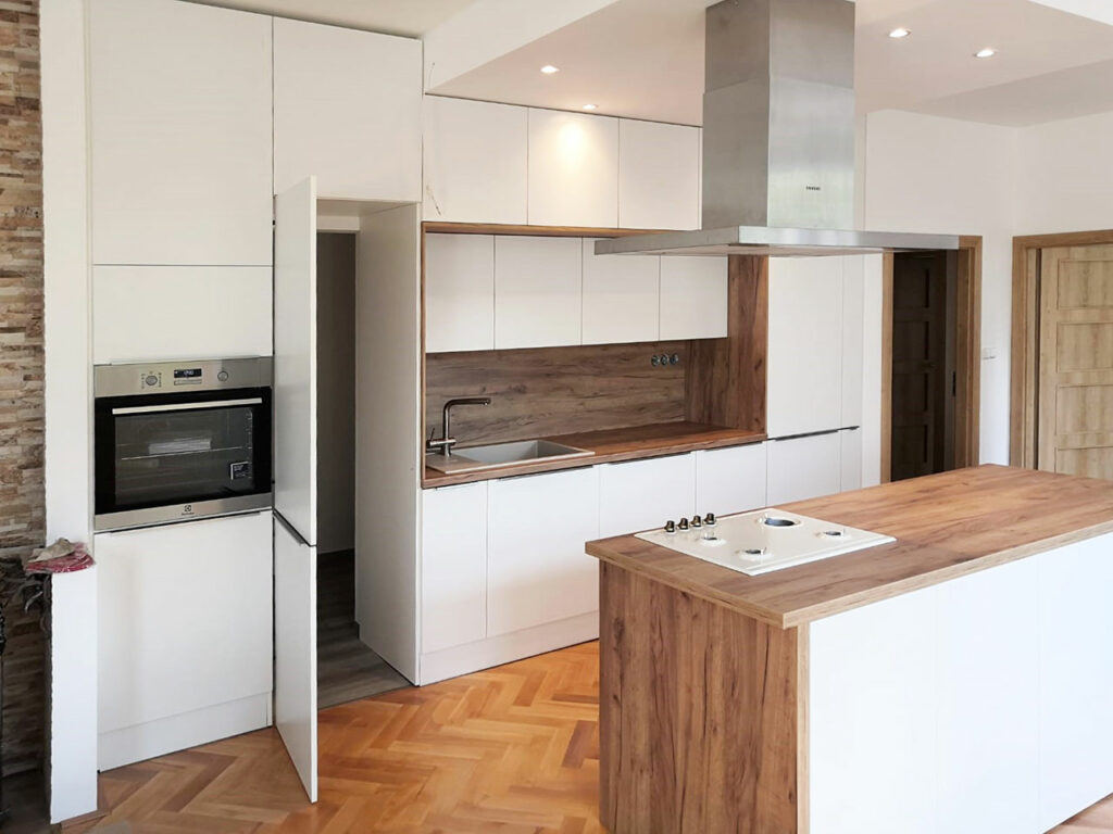 Rovná kcuhyň s ostrůvkem v bílé matné barvě a dubovou pracovní deskou a zástěnou. V kuchyni je skrytý průchod do špajzu.