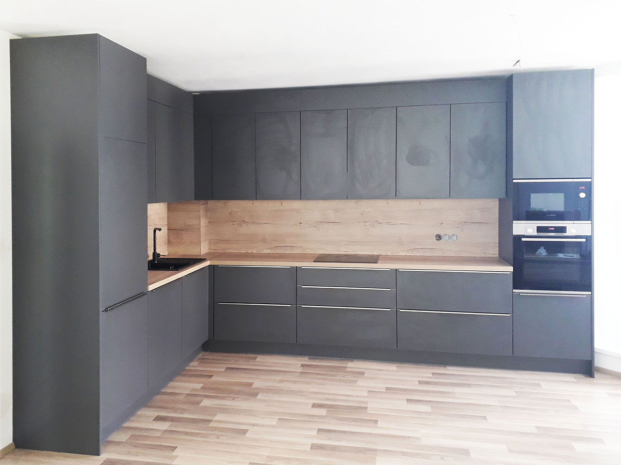 Kuchyň do L v šedé antracitové barvě a dubovou pracovní deskou a zástěnou.